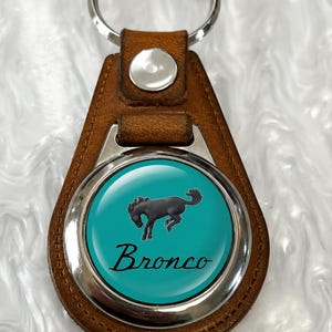 May include: A brown leather keychain featuring a circular metal insert with a glossy teal background. The center displays a black silhouette of a bucking horse above the word Bronco in a black cursive script. A silver metal ring is attached at the top.