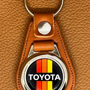 May include: A brown leather keychain with a silver metal charm featuring the Toyota logo with red, orange, and yellow stripes. The logo is on a black background.
