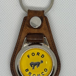 Puede incluir: Llavero de cuero marrón con un anillo de metal plateado y un emblema redondo amarillo con un logotipo Ford Bronco negro.