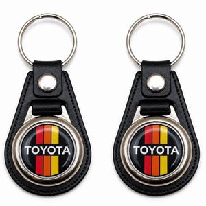 May include: Two black leather keychains with a silver ring and a circular emblem. The emblem features the word "TOYOTA" in white, set against a black background with vertical stripes in red, orange, and yellow.