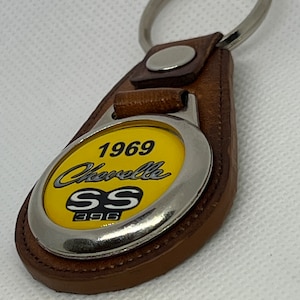 May include: A brown leather keychain with a silver metal charm. The charm features a yellow background with the text "1969 Chevelle SS 396".