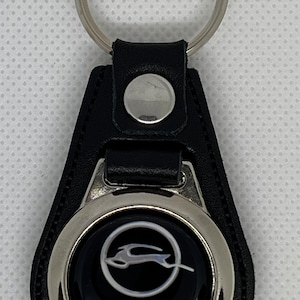 Könnte beinhalten: Schwarzer Leder-Schlüsselanhänger mit einem silbernen Ring und einem runden silbernen Anhänger mit einem weißen Impala-Logo und dem Wort "Impala" in silbernen Buchstaben.