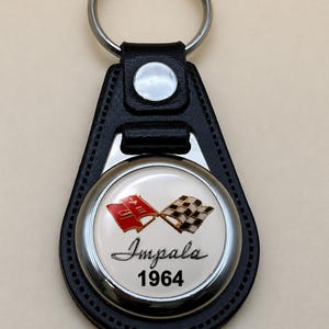 Può includere: Portachiavi in pelle nera con anello argentato ed emblema circolare. L'emblema presenta la scritta "Impala" e l'anno "1964" con due bandiere incrociate rosse e nere e bianche a scacchi.