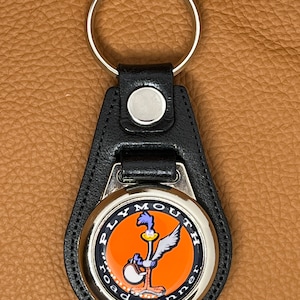 May include: Black leather keychain with a silver round charm featuring the Plymouth Road Runner logo. The logo is a cartoon Road Runner with a bright orange background and the words "Plymouth Road Runner" around the outside.
