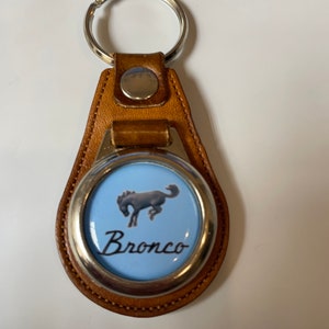 May include: A brown leather keychain with a silver ring and a round metal charm. The charm features a blue background with a white silhouette of a horse and the word "Bronco" in black lettering.