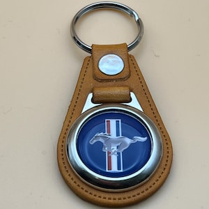 Puede incluir: Un llavero de cuero marrón claro con un llavero plateado. El llavero presenta un emblema circular con fondo azul, un caballo plateado y rayas rojas y blancas. El emblema está en un borde plateado y está fijado en un respaldo de cuero marrón claro.