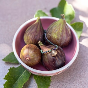 Peut inclure: Un bol rose rempli de figues mûres, certaines avec des rayures vertes et violettes. Un collier de perles avec un pendentif en forme de cœur repose sur une figue. Des feuilles de figuier vertes entourent le bol.