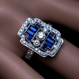 May include: A silver ring with a large, rectangular, blue sapphire surrounded by smaller diamonds. The ring has a geometric design and is set in a white metal.