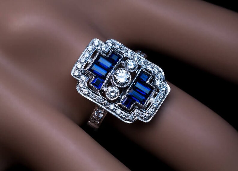 May include: A silver ring with a large, rectangular, blue sapphire surrounded by smaller diamonds. The ring has a geometric design and is set in a white metal.