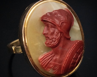 May include: A gold ring featuring an oval cameo with a carved profile of a man in a helmet. The cameo is red and set against a cream-colored background. The ring has a classic design.