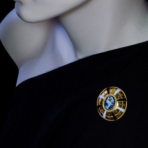 May include: A gold-toned brooch with a white and black enamel design. The center of the brooch features a cameo with a portrait of a woman.