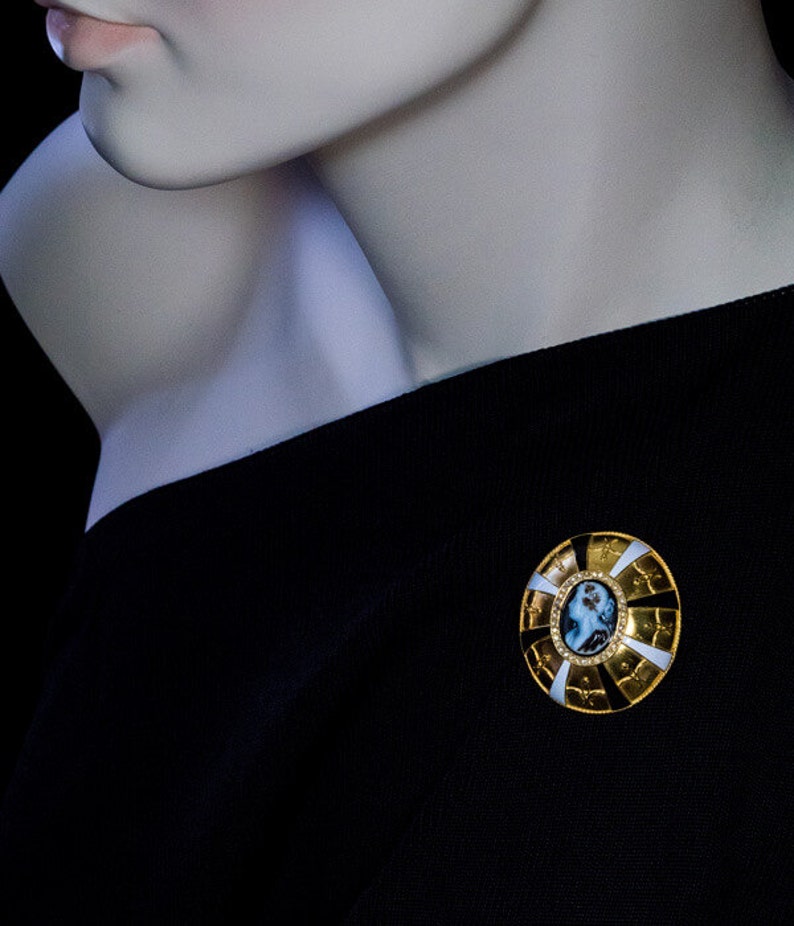 May include: A gold-toned brooch with a white and black enamel design. The center of the brooch features a cameo with a portrait of a woman.