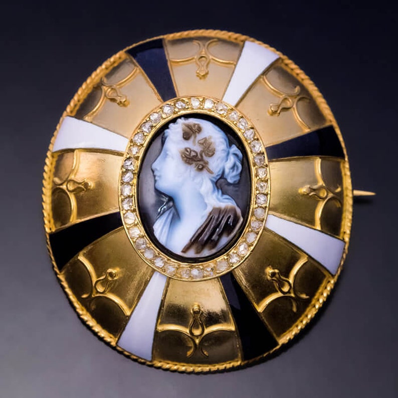 May include: A gold brooch with a cameo of a woman's profile set in a circle of diamonds. The brooch is decorated with black and white enamel and gold accents.
