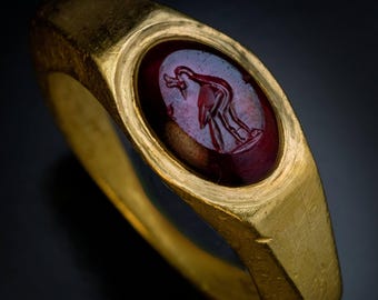 May include: A gold ring featuring an oval, deep red gemstone. The stone is intricately carved with a bird motif. The ring has a rectangular band and a polished surface. The ring is displayed against a dark backdrop.