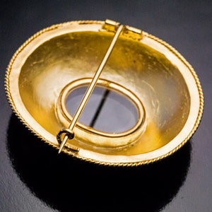 May include: A gold-toned brooch with a round, slightly domed design. The brooch has a circular opening in the center, revealing a blue stone.