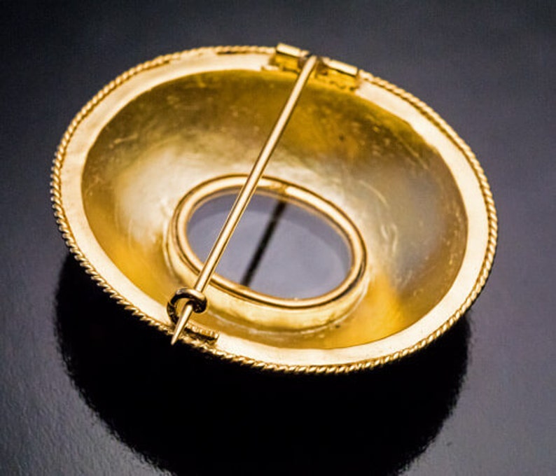 May include: A gold-toned brooch with a round, slightly domed design. The brooch has a circular opening in the center, revealing a blue stone.