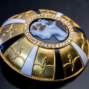 May include: A gold and enamel brooch with a cameo portrait of a woman. The cameo is surrounded by a circle of diamonds. The brooch is decorated with black and white enamel.