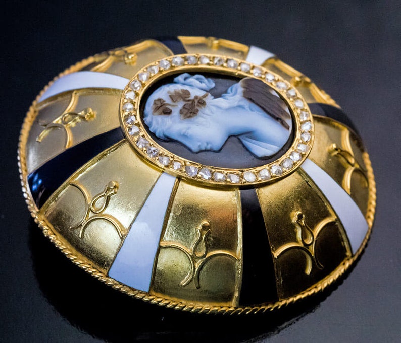 May include: A gold and enamel brooch with a cameo portrait of a woman. The cameo is surrounded by a circle of diamonds. The brooch is decorated with black and white enamel.