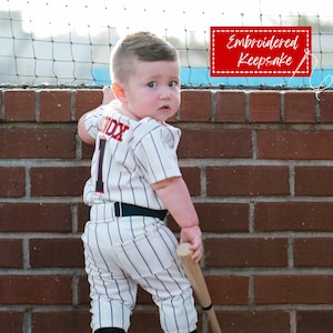 May include: A baby wearing a baseball uniform with vertical blue stripes, the number 1, and the name "SOX" on the back. The uniform includes a belt and a wooden baseball bat. The image also includes the text "Embroidered Keepsake" in a red box.