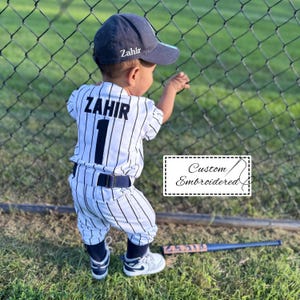 May include: A toddler wearing a custom baseball uniform with the name "Zahir" and the number "1" on the back. The uniform is white with black pinstripes and a navy blue belt. A navy blue cap with "Zahir" is worn.
