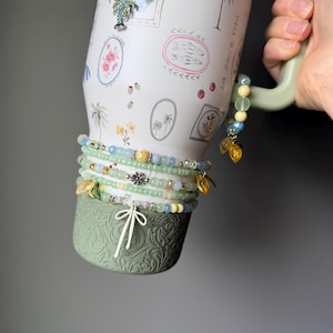 May include: A white travel mug with a green handle and base, adorned with a tropical design and the words "La Dolce Vita." Several beaded bracelets in shades of green, blue, and yellow are wrapped around the mug, some with charm accents.