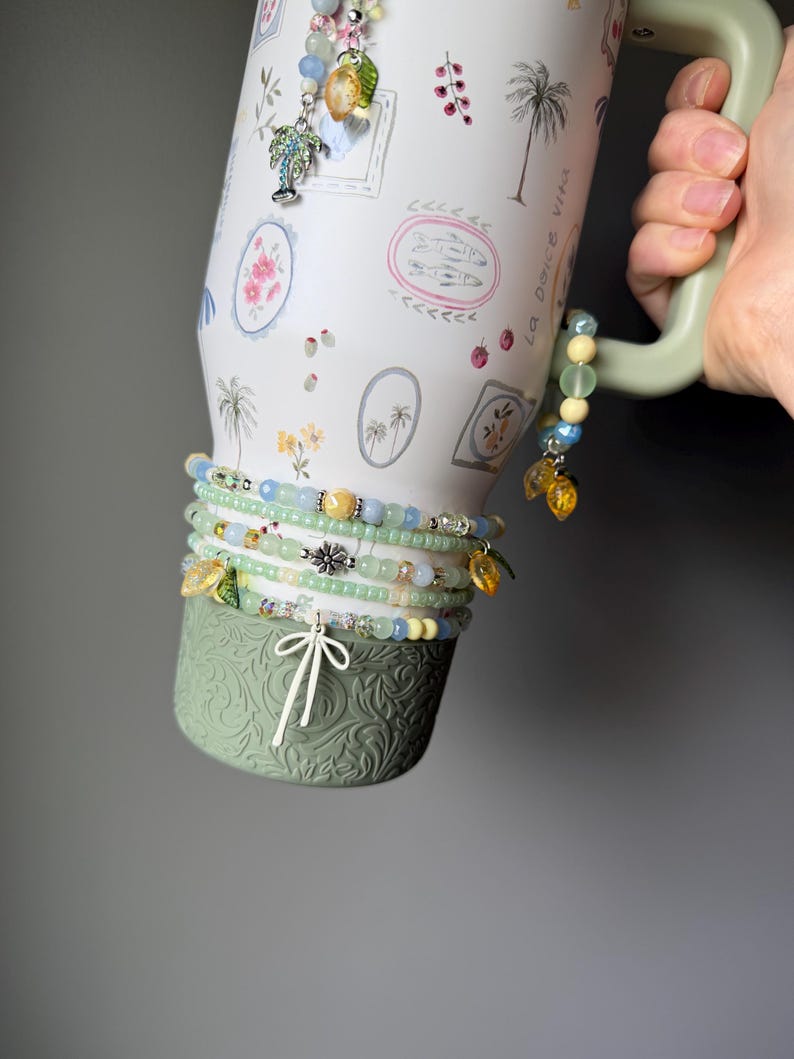 May include: A white travel mug with a green handle and base, adorned with a tropical design and the words "La Dolce Vita." Several beaded bracelets in shades of green, blue, and yellow are wrapped around the mug, some with charm accents.