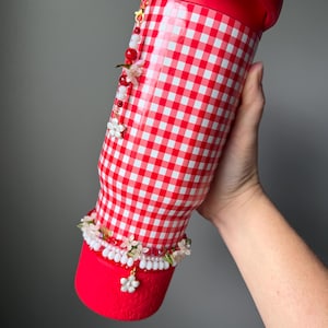 K&ouml;nnte beinhalten: Ein rot-wei&szlig; karierter Becher mit rotem Deckel und Boden. Der Becher ist mit einem floralen und perlenbesetzten Anh&auml;nger und einem dekorativen Blumenband verziert. Der Becher wird von einer Person gehalten.