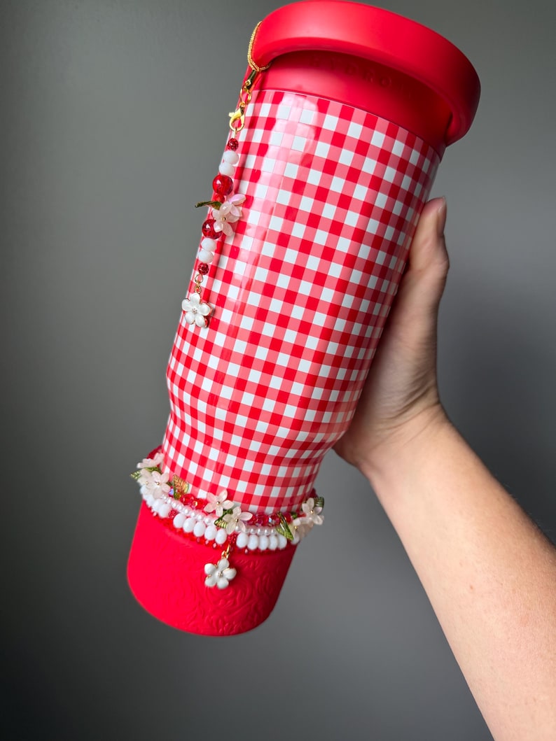 K&ouml;nnte beinhalten: Ein rot-wei&szlig; karierter Becher mit rotem Deckel und Boden. Der Becher ist mit einem floralen und perlenbesetzten Anh&auml;nger und einem dekorativen Blumenband verziert. Der Becher wird von einer Person gehalten.
