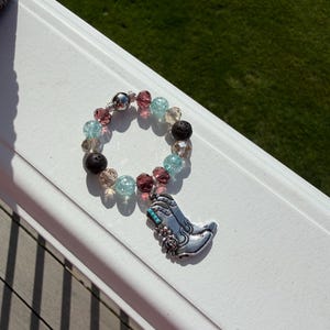 Puede incluir: Una pulsera de cuentas con un dije de bota de vaquero plateada. La pulsera presenta una mezcla de cuentas transparentes, rosas, azules y negras. El dije de bota de vaquero tiene detalles turquesa y florales.