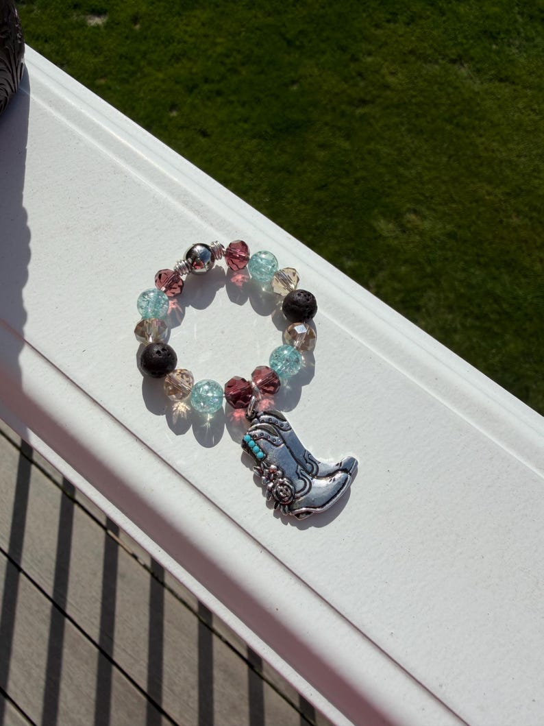 Puede incluir: Una pulsera de cuentas con un dije de bota de vaquero plateada. La pulsera presenta una mezcla de cuentas transparentes, rosas, azules y negras. El dije de bota de vaquero tiene detalles turquesa y florales.
