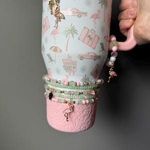 Puede incluir: Un vaso blanco con asa rosa que presenta un patr&oacute;n tropical de flamencos rosas, palmeras, coches antiguos y letreros de motel. El vaso est&aacute; decorado con varias pulseras de cuentas en tonos rosa y verde, con peque&ntilde;os dijes de flamencos dorados y cuentas colgantes.