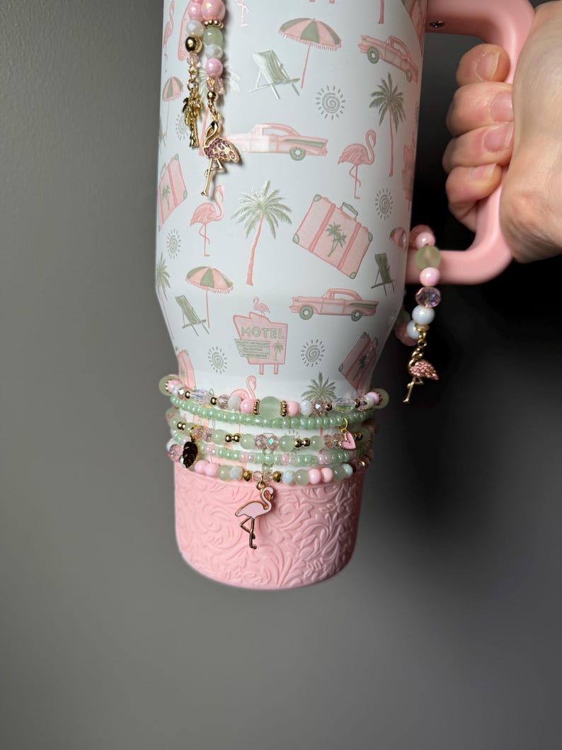 Puede incluir: Un vaso blanco con asa rosa que presenta un patr&oacute;n tropical de flamencos rosas, palmeras, coches antiguos y letreros de motel. El vaso est&aacute; decorado con varias pulseras de cuentas en tonos rosa y verde, con peque&ntilde;os dijes de flamencos dorados y cuentas colgantes.
