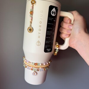 May include: A cream-colored 40oz HydroJug tumbler with a handle, decorated with beaded charms featuring sunflower and daisy motifs. The tumbler has a black label with white text listing features like cup holder friendly and leak proof.