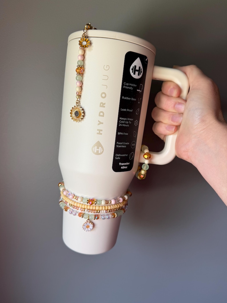 May include: A cream-colored 40oz HydroJug tumbler with a handle, decorated with beaded charms featuring sunflower and daisy motifs. The tumbler has a black label with white text listing features like cup holder friendly and leak proof.