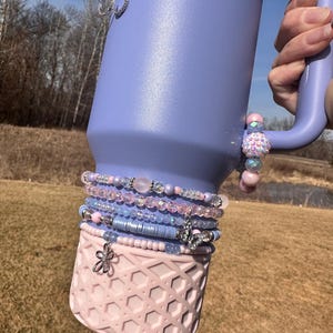 May include: A purple stainless steel tumbler with a pink silicone lid and a butterfly charm. The tumbler is decorated with multiple layers of beaded bracelets in shades of pink, blue, and white.