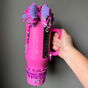 May include: A pink HydroJug water bottle with a large, glittery purple and pink bow on top. The bottle is adorned with beaded accents in shades of pink and purple. The handle is held by a hand.