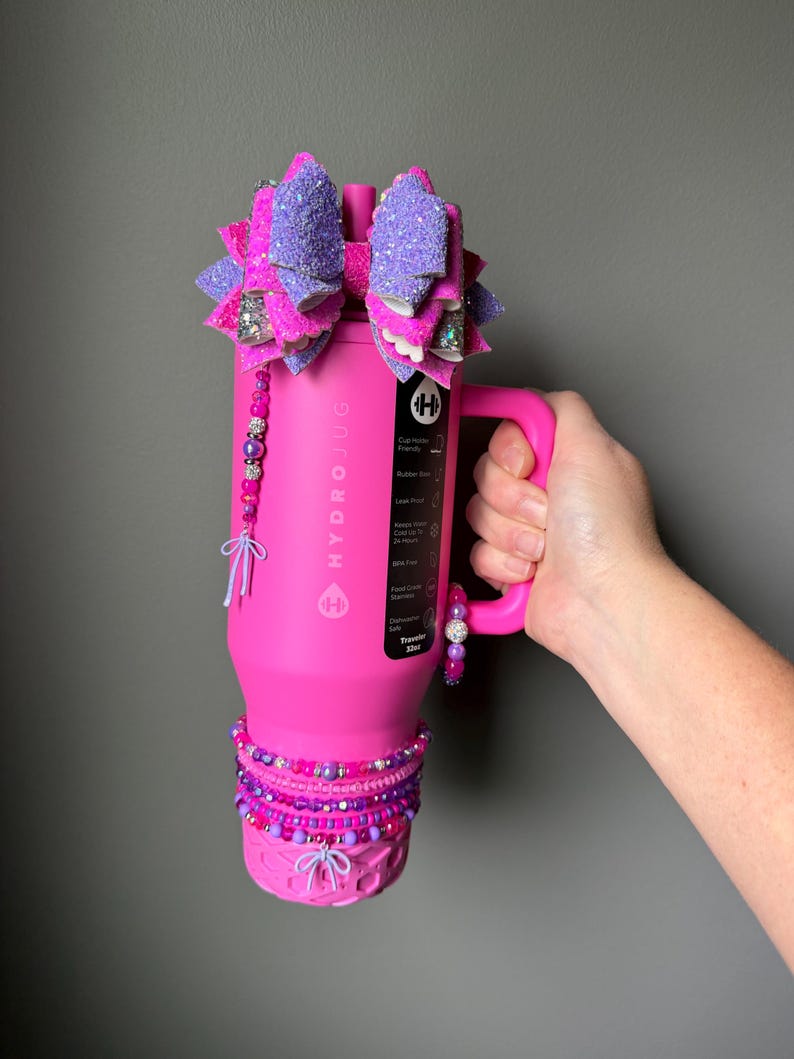 May include: A pink HydroJug water bottle with a large, glittery purple and pink bow on top. The bottle is adorned with beaded accents in shades of pink and purple. The handle is held by a hand.