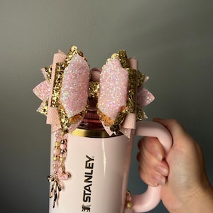 Könnte beinhalten: Ein hellrosa Stanley-Trinkbecher mit einer großen dekorativen Schleife aus geschichtetem rosa und goldfarbenem Glitzerstoff an der Oberseite. Ein Perlenanhänger mit kleinen rosa und goldenen Perlen sowie einem herzförmigen Anhänger hängt an der Seite.