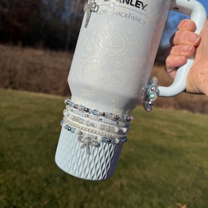 May include: A light blue Stanley tumbler with a white floral pattern and the text "STANLEY LoveHackFancy". The tumbler is adorned with beaded bracelets, a bow charm, and a cross charm. The handle is white.