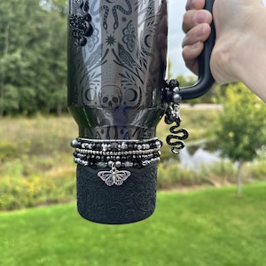 May include: A black Stanley tumbler with intricate etched designs, including a skull, snakes, and celestial motifs. The tumbler is adorned with beaded charms and a decorative butterfly charm. The handle is black, and the base has a textured, patterned sleeve.