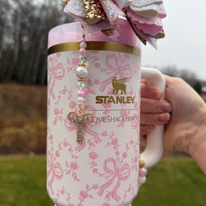 May include: A white and pink floral Stanley mug with a gold rim and handle. The mug features the "LOVESHACKFANCY" logo and a decorative cross charm. A large pink and gold bow adorns the top, and a beaded bracelet wraps around the base.