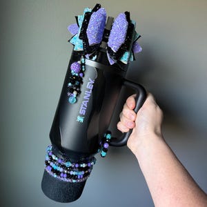 May include: Black Stanley tumbler with a handle, adorned with purple glitter bows, beaded accents, and a decorative base. The tumbler features the Stanley logo and is embellished with various shades of purple, black, and teal beads.