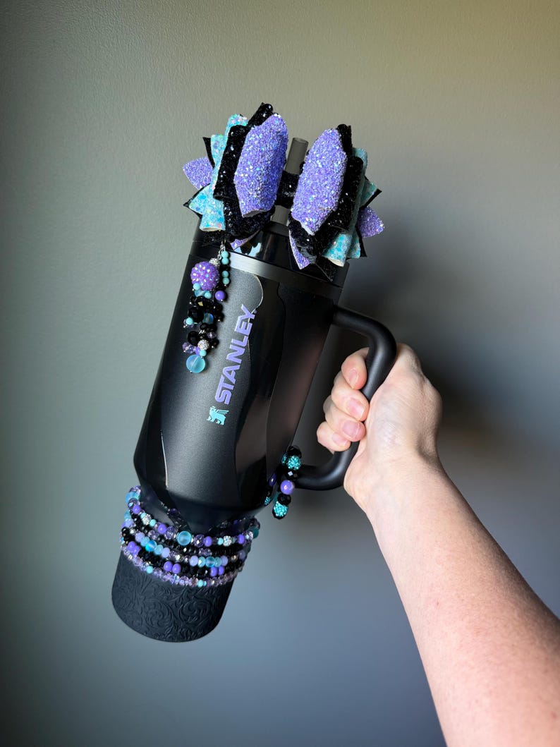 May include: Black Stanley tumbler with a handle, adorned with purple glitter bows, beaded accents, and a decorative base. The tumbler features the Stanley logo and is embellished with various shades of purple, black, and teal beads.