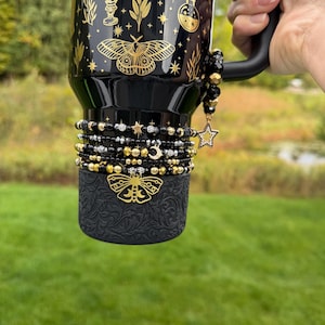 May include: A black tumbler featuring gold celestial and moth illustrations, adorned with a beaded bracelet stack in black, gold, and silver tones. The bracelet includes a dangling star charm and a gold moth charm, with a textured black base.
