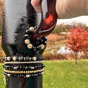 May include: A stack of black and gold beaded bracelets on a black textured surface. The bracelets feature various bead sizes and gold accents. The background shows a blurred outdoor scene with trees and grass.