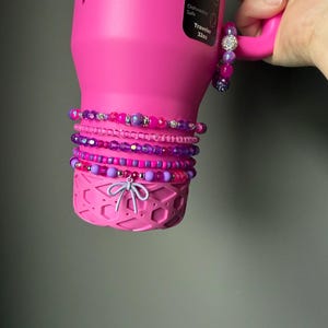 May include: A pink insulated water bottle with a handle, adorned with multiple beaded bracelets in shades of pink and purple. The bracelets feature various bead sizes and textures, including crystal accents. The bottle has a decorative bow at the base.