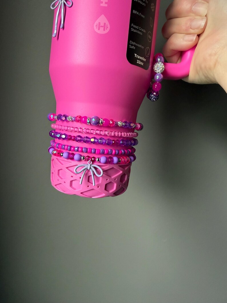 May include: A pink insulated water bottle with a handle, adorned with multiple beaded bracelets in shades of pink and purple. The bracelets feature various bead sizes and textures, including crystal accents. The bottle has a decorative bow at the base.