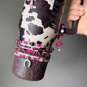 May include: A 40oz stainless steel tumbler with a brown and cream cow print pattern, featuring a dark brown base with an embossed floral design. The tumbler is adorned with multiple beaded bracelets in shades of pink, brown, and silver, including charms shaped like a cowboy boot, a cowboy hat, and a horseshoe.