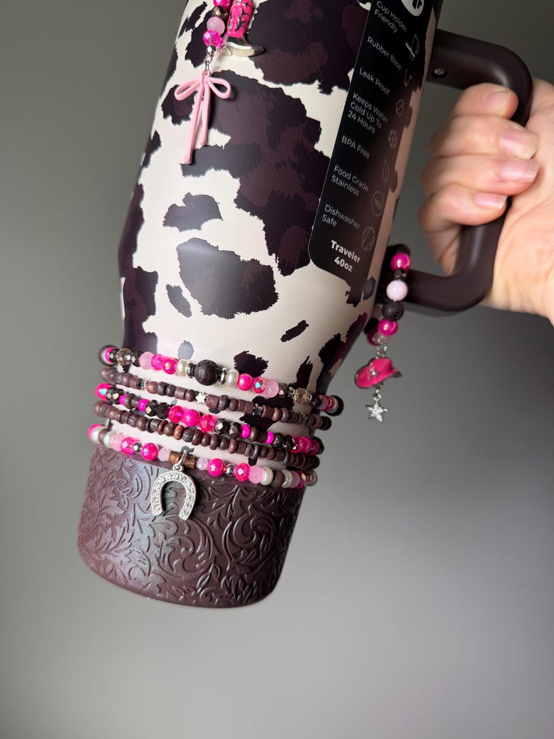 May include: A 40oz stainless steel tumbler with a brown and cream cow print pattern, featuring a dark brown base with an embossed floral design. The tumbler is adorned with multiple beaded bracelets in shades of pink, brown, and silver, including charms shaped like a cowboy boot, a cowboy hat, and a horseshoe.
