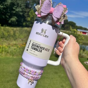K&ouml;nnte beinhalten: Ein wei&szlig;er Stanley 40 oz (1,18 L) "The Flowstate" Quencher H2.0 Tumbler mit einer Schleife aus lila und goldenen Glitzer oben. Der Becher hat einen Griff und einen Strohhalm. Er ist mit einer Reihe von lila und rosa Perlenarmb&auml;ndern verziert.